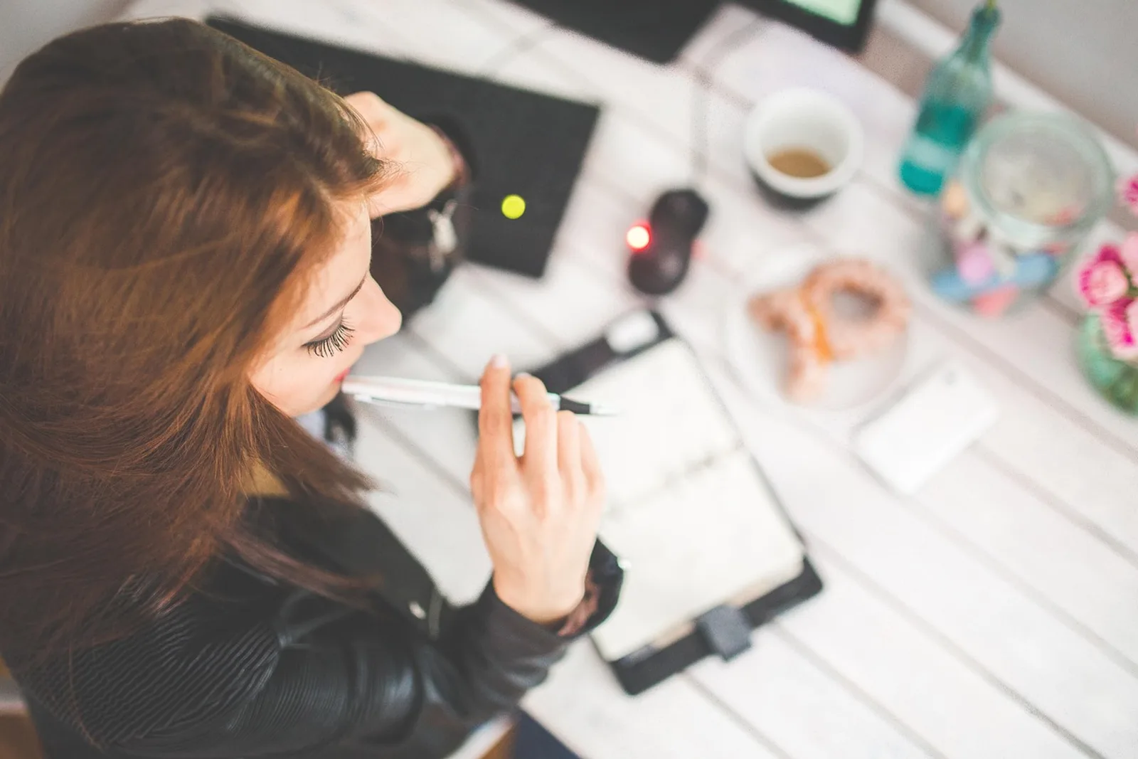 Woman holding a pen to her lips above her notebook at her desk, wondering why she never feels good enough