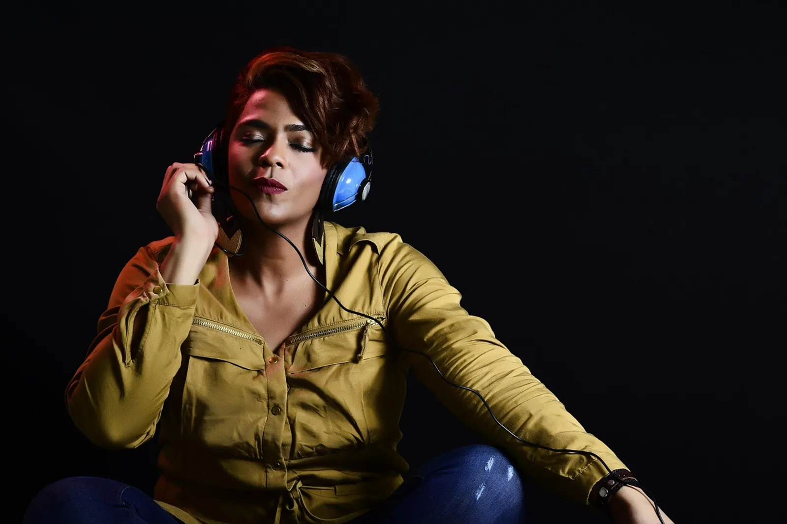 Woman with closed eyes and blue headphones sitting against a dark background, listening to a hypnosis recording