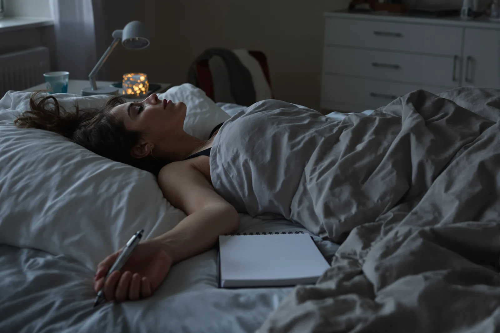 Frau liegt abends mit geschlossenen Augen im Bett, Stift und Notizbuch neben sich, Ruhemoment zur posthypnotischen Suggestion