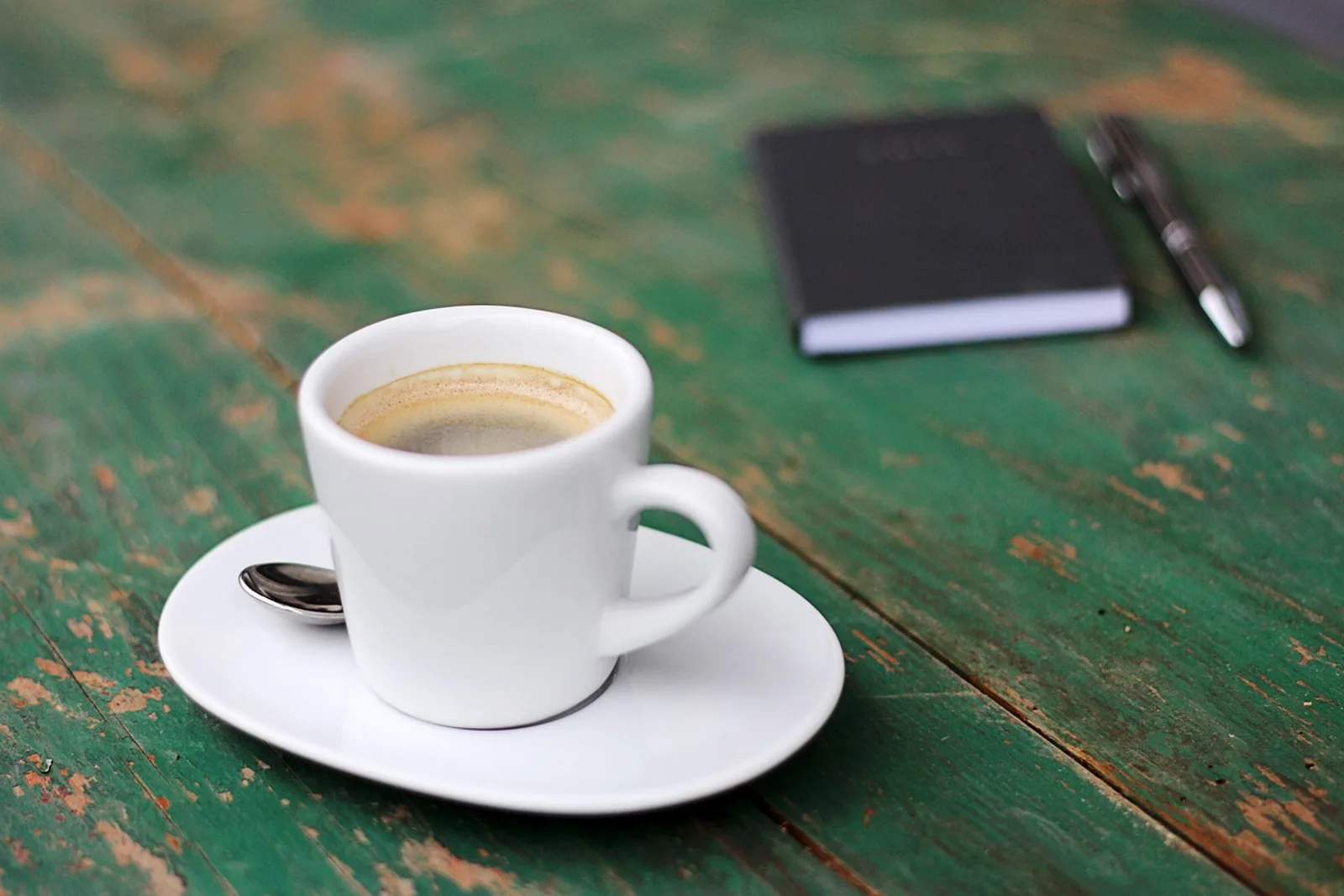 Espresso on a green wooden table with a closed notebook and pen behind it, ready to prioritize tasks in the morning