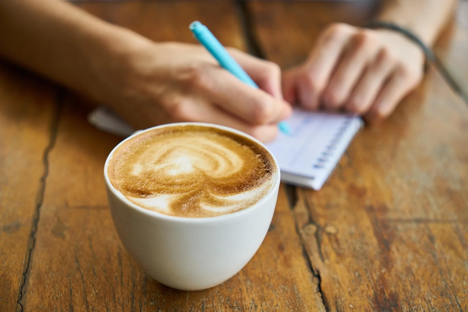 Hände notieren mit hellblauem Stift einen Satz, daneben ein Cappuccino – kleines Ritual gegen den inneren Kritiker